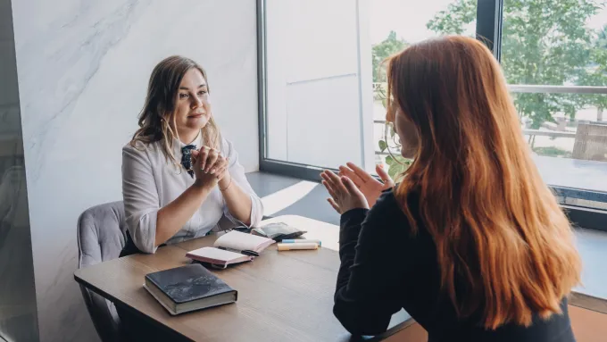 Two women talking sitting at a desk by a window