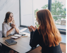 Two women talking sitting at a desk by a window